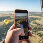 A person in a hot air balloon holds a smartphone, capturing a photo of two other hot air balloons floating over a scenic landscape.