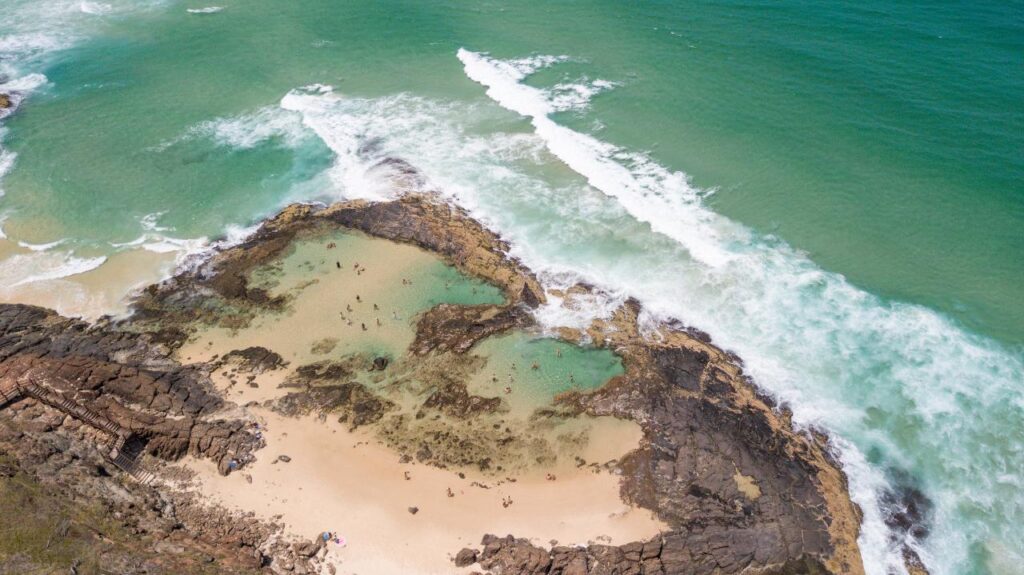 Aerial view of a rocky coastline with small tidal pools, sandy beach, and turquoise waves breaking along the shore.