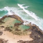 Aerial view of a rocky coastline with small tidal pools, sandy beach, and turquoise waves breaking along the shore.
