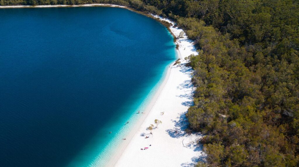 Aerial view of a lake with deep blue water, a white sandy shoreline, and dense green forest surrounding the area. A few people are visible on the beach.