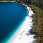 Aerial view of a lake with deep blue water, a white sandy shoreline, and dense green forest surrounding the area. A few people are visible on the beach.