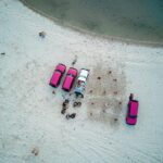 Aerial view of four vehicles parked on a sandy beach near water, with groups of people gathered around them.