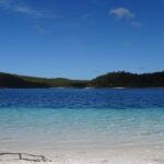 A clear lake with blue water and white sandy shore, surrounded by green forest under a sunny blue sky.