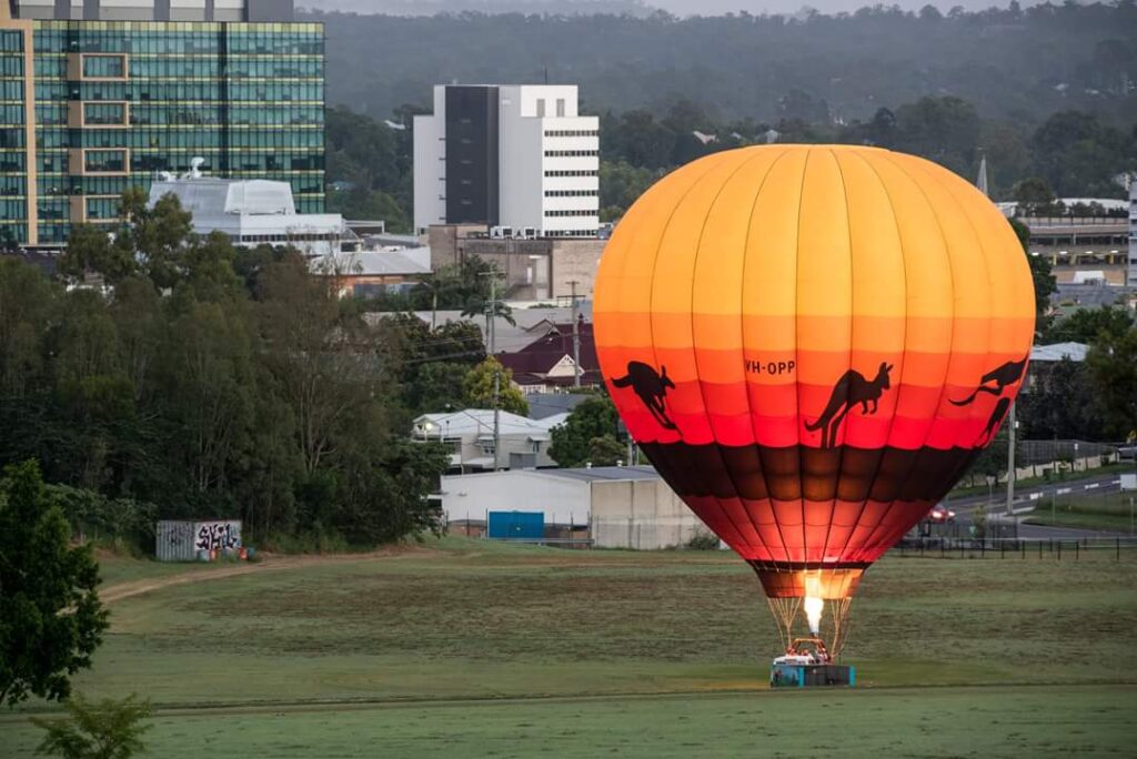 A hot air balloon with kangaroo silhouettes is grounded on a grassy field with city buildings and trees in the background.