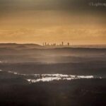 A distant city skyline rises above layers of misty hills and reflective water under a cloudy, golden sky at sunrise or sunset.