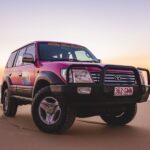 A red Toyota SUV with off-road tires is parked on a sandy beach at sunset, with the ocean and pastel sky in the background.
