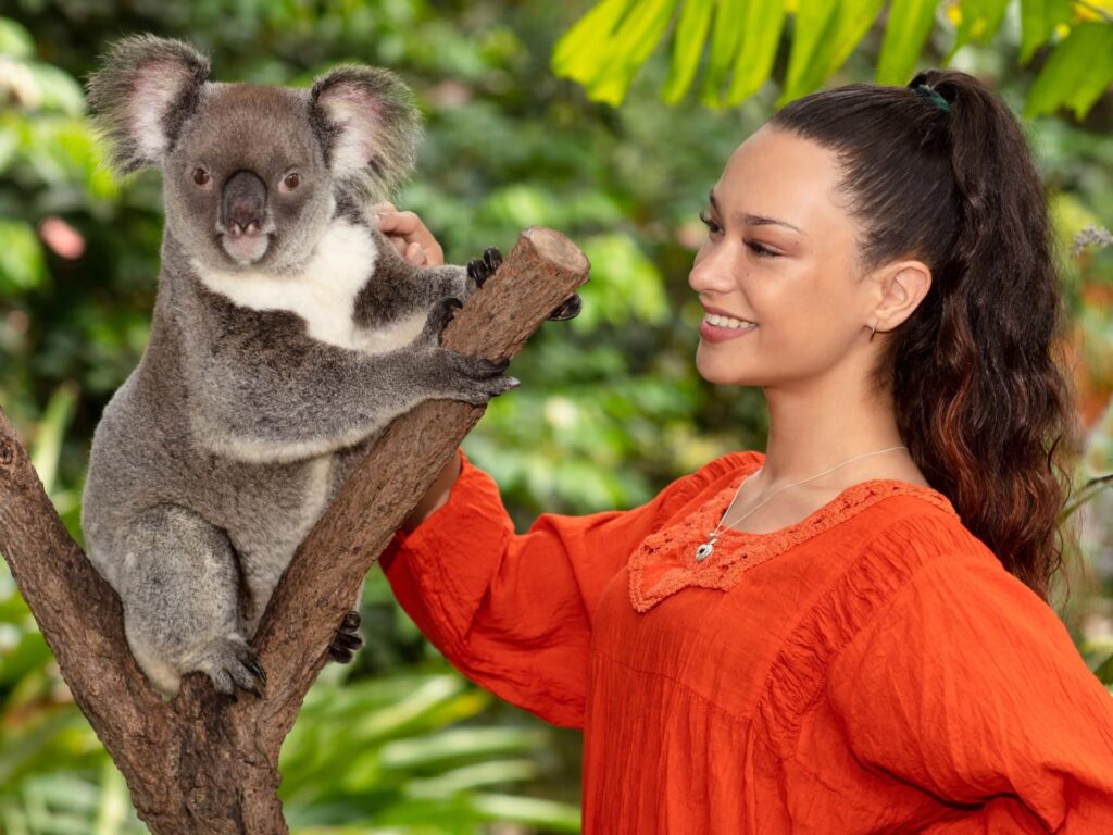 A woman in an orange top stands next to a koala perched on a tree branch, both set against a lush, green natural background.