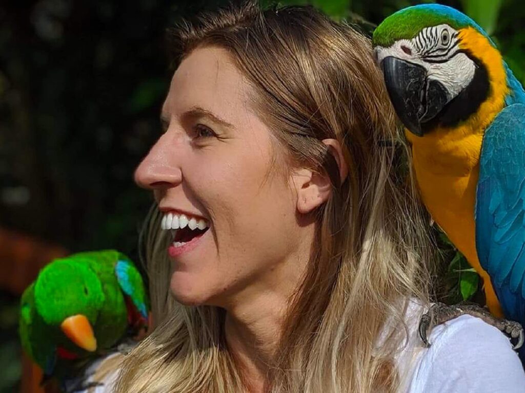 A woman with long blonde hair smiles while two colorful parrots perch on her shoulders, one green and one blue and yellow.