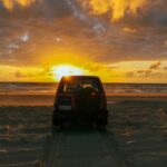A red SUV parked on sandy beach tire tracks faces the ocean at sunset, with dramatic clouds and golden sunlight in the sky.