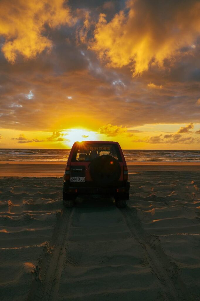 A red SUV parked on sandy beach tire tracks faces the ocean at sunset, with dramatic clouds and golden sunlight in the sky.