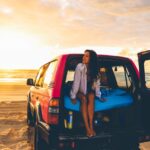A woman sits on the open tailgate of a red SUV parked on a sandy beach at sunset, with the ocean and sky in the background.
