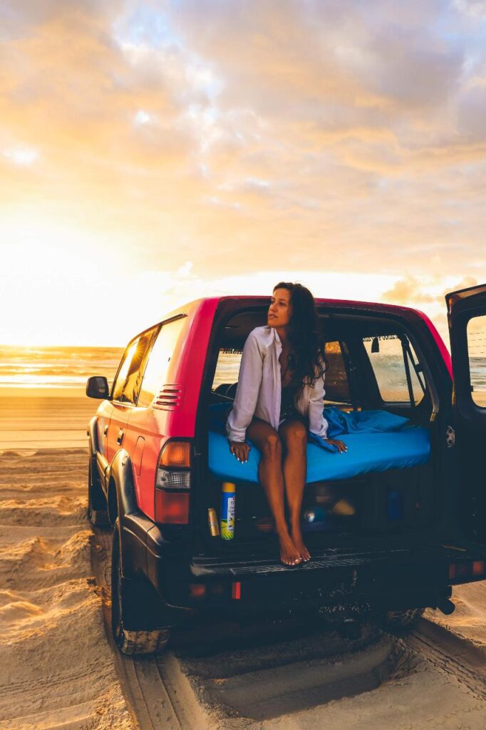 A woman sits on the open tailgate of a red SUV parked on a sandy beach at sunset, with the ocean and sky in the background.