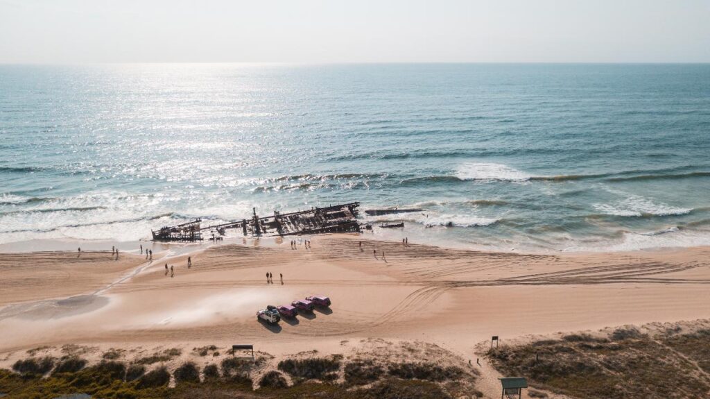 Aerial view of a beach with people walking near a large, rusted shipwreck partially submerged in the ocean and several vehicles parked on the sand.