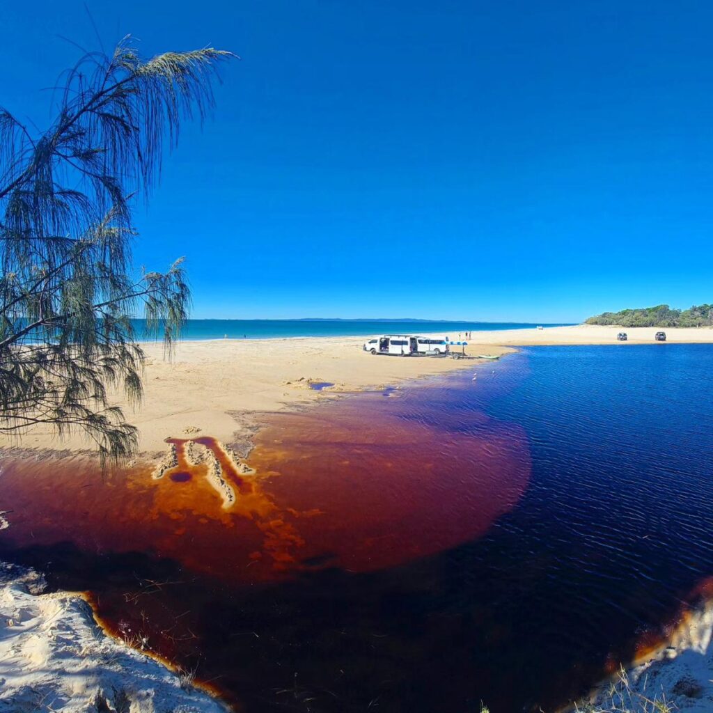 A sandy beach with a reddish-brown lagoon meeting the ocean, a few vehicles and people on the shore, and clear blue sky overhead.