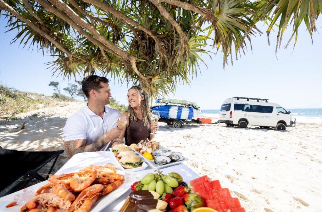 A man and woman sit at a table on a beach, enjoying seafood and fruit, with a van and surfboards parked nearby under a tree.