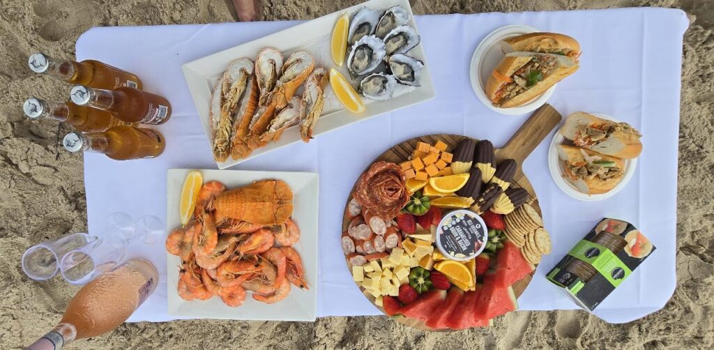 A picnic spread on a white cloth with seafood, charcuterie board, sandwiches, beer bottles, wine, and plastic cups set on sand.