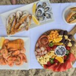 A picnic spread on a white cloth with seafood, charcuterie board, sandwiches, beer bottles, wine, and plastic cups set on sand.