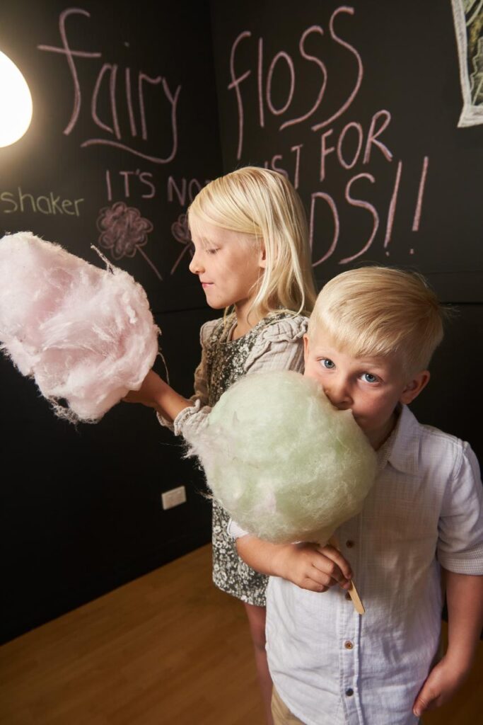 Two young children holding and eating large sticks of cotton candy, standing in front of a chalkboard wall with "fairy floss" written on it.