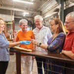 A woman wearing gloves hands an object to a group of four smiling adults standing by a counter in an industrial indoor setting.