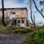 An old concrete building with barred windows and doors stands amid trees and greenery; three people are visible in the distance near the ocean.
