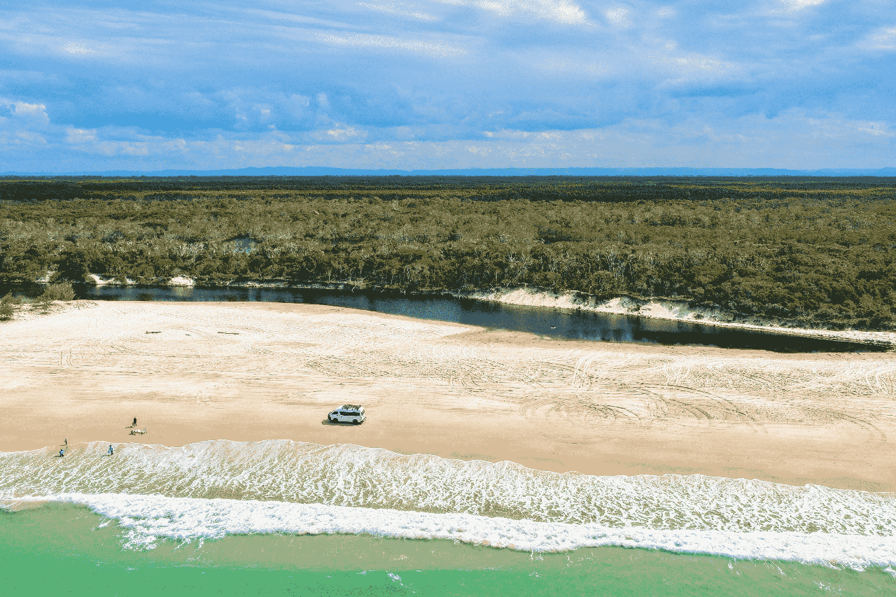 Aerial view of a sandy beach with a parked vehicle near the shoreline, a few people nearby, a narrow river, forest in the background, and waves approaching the shore.