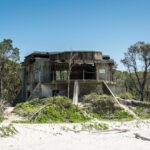 An abandoned concrete bunker stands on a sandy beach, surrounded by sparse vegetation and trees under a clear blue sky.