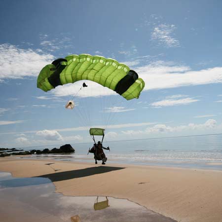 A person with a green parachute is landing on a sandy beach near the shoreline under a blue sky with scattered clouds.