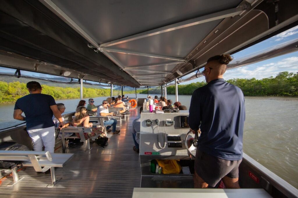 People seated on benches and standing aboard a covered river tour boat, traveling along a scenic river bordered by greenery under a clear sky.