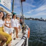 A group of people sits on the edge of a boat, smiling and enjoying a sunny day on the water near a city shoreline.