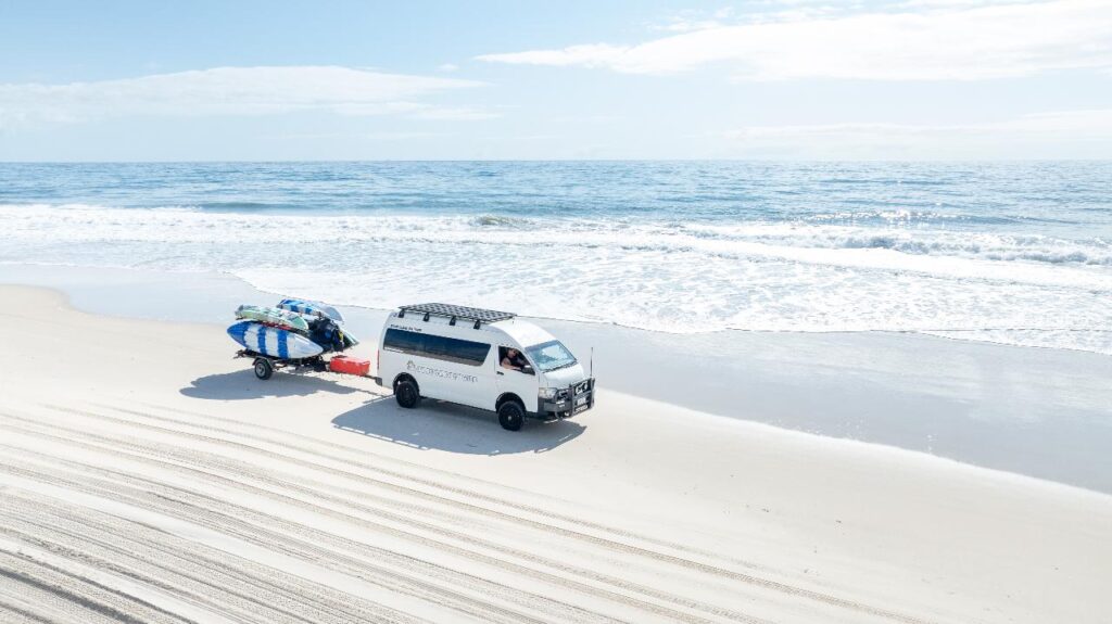 A white van towing a trailer with surfboards is parked on a sandy beach near the ocean with waves in the background.