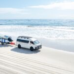 A white van towing a trailer with surfboards is parked on a sandy beach near the ocean with waves in the background.