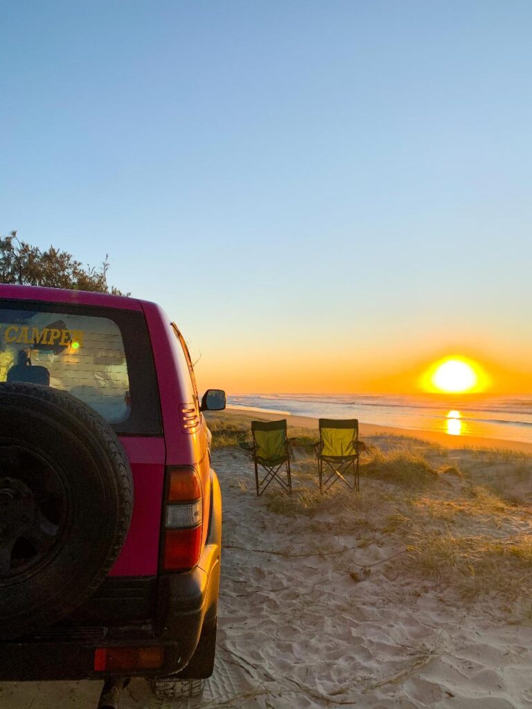 A red SUV parked on sandy ground near two empty camping chairs facing the ocean at sunset, with clear skies and the sun low on the horizon.