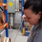 A person draws liquid from a wooden barrel using a thief in a storage area lined with barrels on shelves.