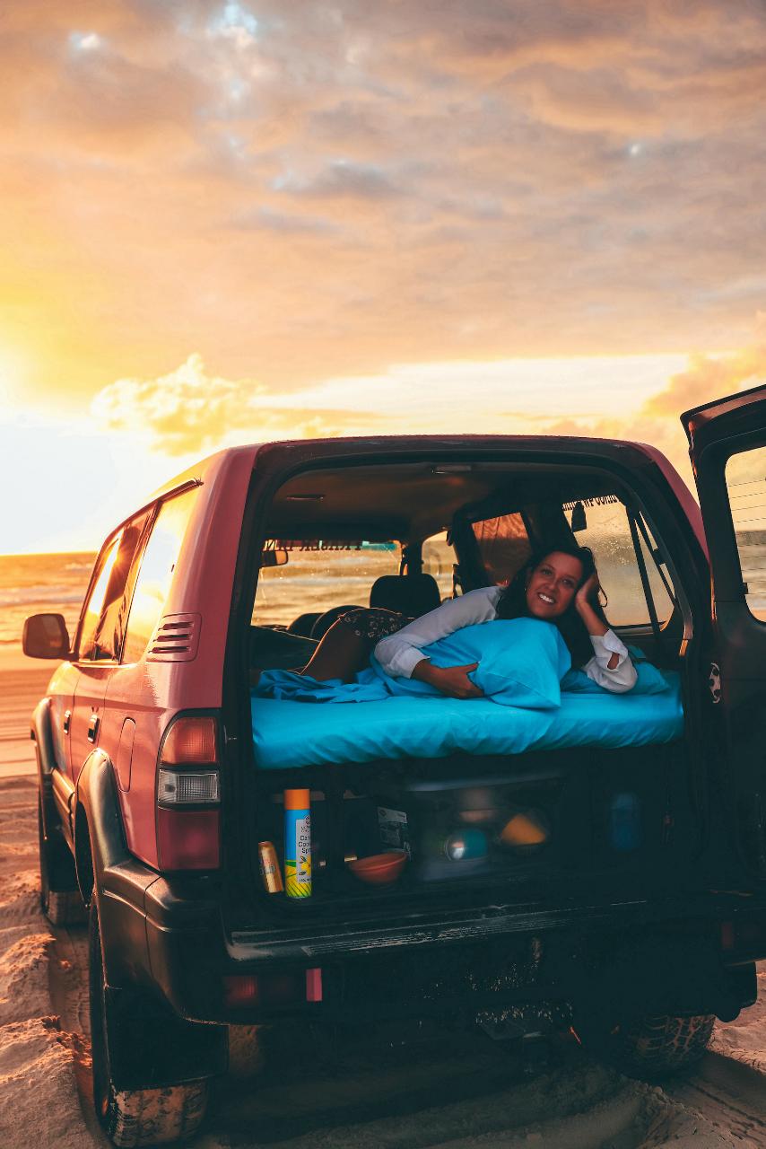 A person lies on a mattress in the back of an SUV parked on a sandy beach at sunset, with the vehicle’s rear and side doors open.