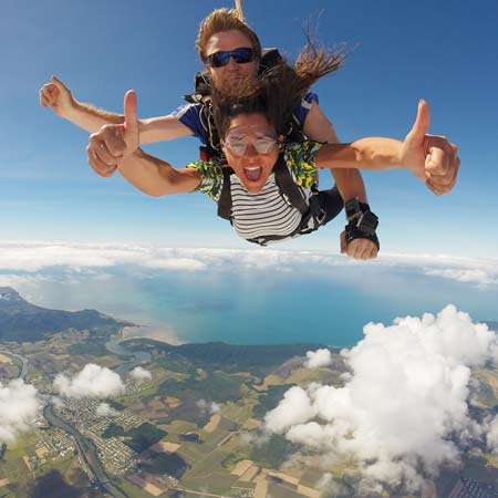 Two people tandem skydiving above clouds and landscape, both smiling and giving thumbs up, with a clear view of land, water, and sky below.
