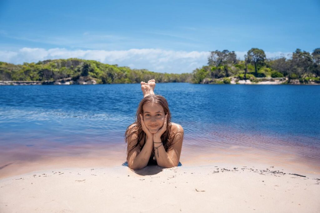 A young girl lies on her stomach at the edge of a sandy beach, resting her chin on her hands, with a blue lake and green trees in the background under a clear sky.