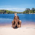 A young girl lies on her stomach at the edge of a sandy beach, resting her chin on her hands, with a blue lake and green trees in the background under a clear sky.