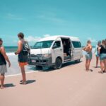 A group of people stand on a sandy beach near a white van with its door open, parked close to the shoreline under a clear blue sky.