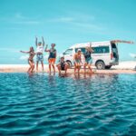 Six people in swimwear pose and jump excitedly by the water on a sandy beach, with a white camper van parked in the background under a clear blue sky.