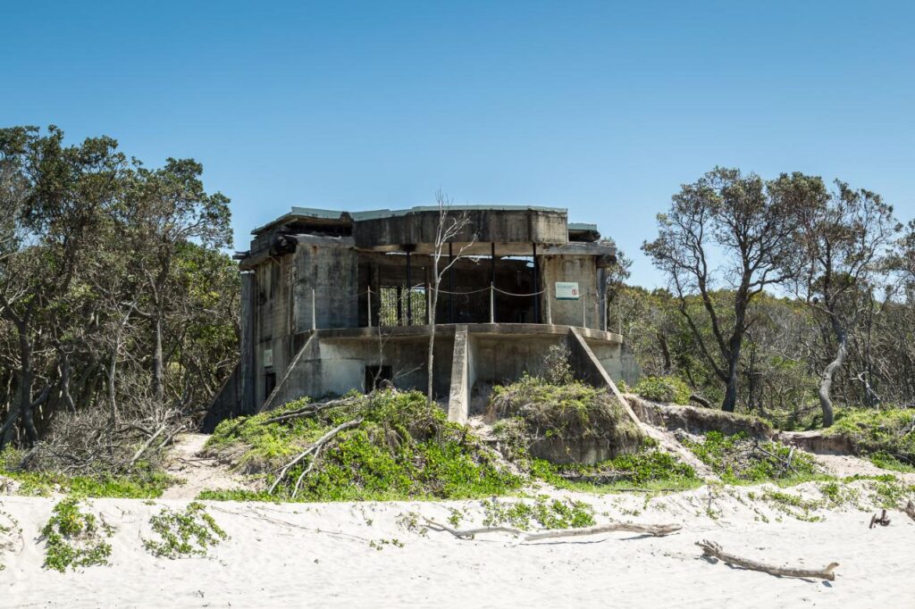 Abandoned concrete bunker with broken windows and overgrown vegetation, situated on a sandy beach with trees in the background under a clear blue sky.