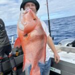 Person on a boat holding a large red fish with the ocean and fishing gear visible in the background.
