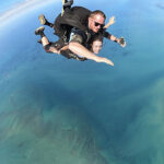 Two people tandem skydiving high above blue water, with their arms outstretched, captured mid-air against a clear sky.