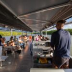 A group of people sit and stand on a covered riverboat as it travels along a river surrounded by greenery under a sunny sky.