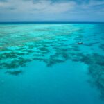 Aerial view of a clear turquoise ocean with patches of coral reefs and a small boat floating on the water under a partly cloudy sky.