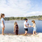 Four people, two adults and two children, stand by a sandy lakeshore under a partly cloudy sky with trees in the background. One adult helps a child up the sandbank.