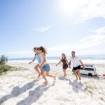 A family of four walks barefoot up a sandy beach dune on a sunny day, with a parked van and surfboards visible in the background near the shoreline.