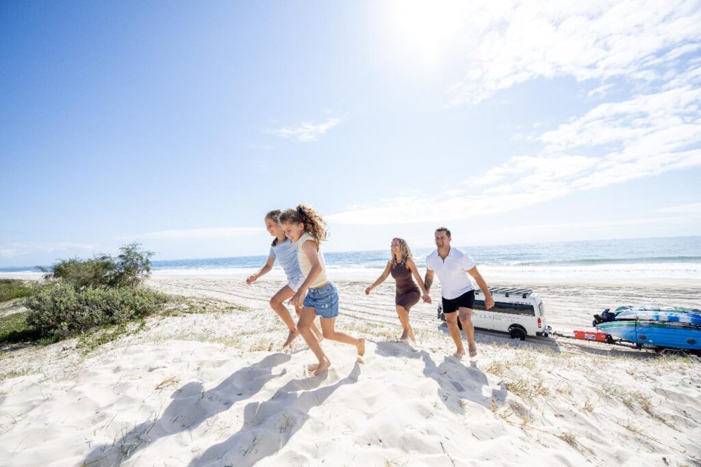 Four people walk up a sandy dune at the beach on a sunny day, with the ocean, a van, and surfboards visible in the background.