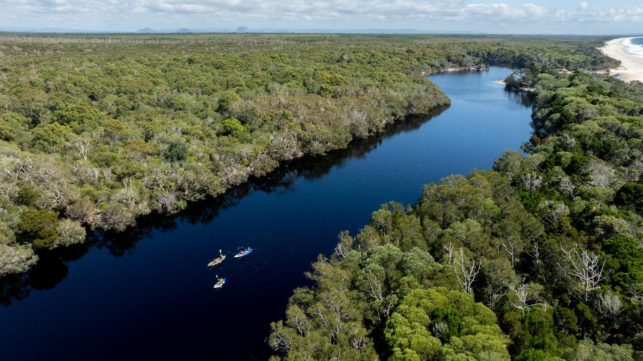 Aerial view of three kayaks on a winding river surrounded by dense forest, with a sandy beach and ocean visible in the distance.