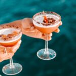 Two hands hold cocktail glasses with frothy drinks, garnished with dried citrus slices and a rim of what appears to be chocolate, against a blue water background.