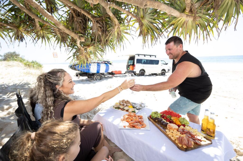 A man and woman exchange drinks at a picnic table set with seafood, fruit, and snacks on a sandy beach under leafy trees, with vans and surfboards in the background.
